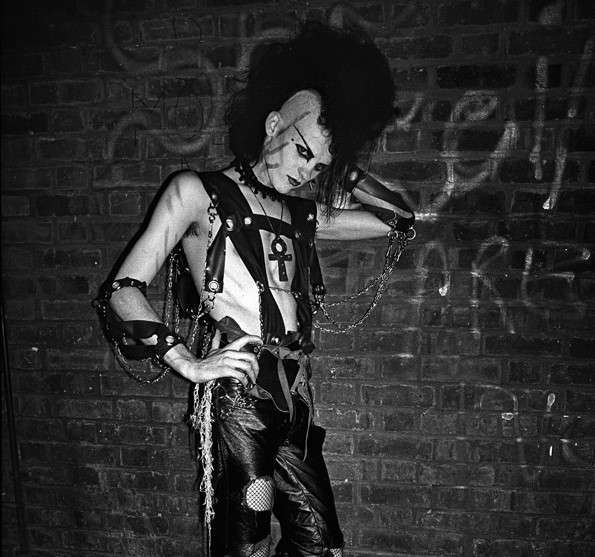 Black and white photo of a Batcave-era goth with a mohawk, harness, leather pants, and Egyptian ankh necklace, standing against a brick wall