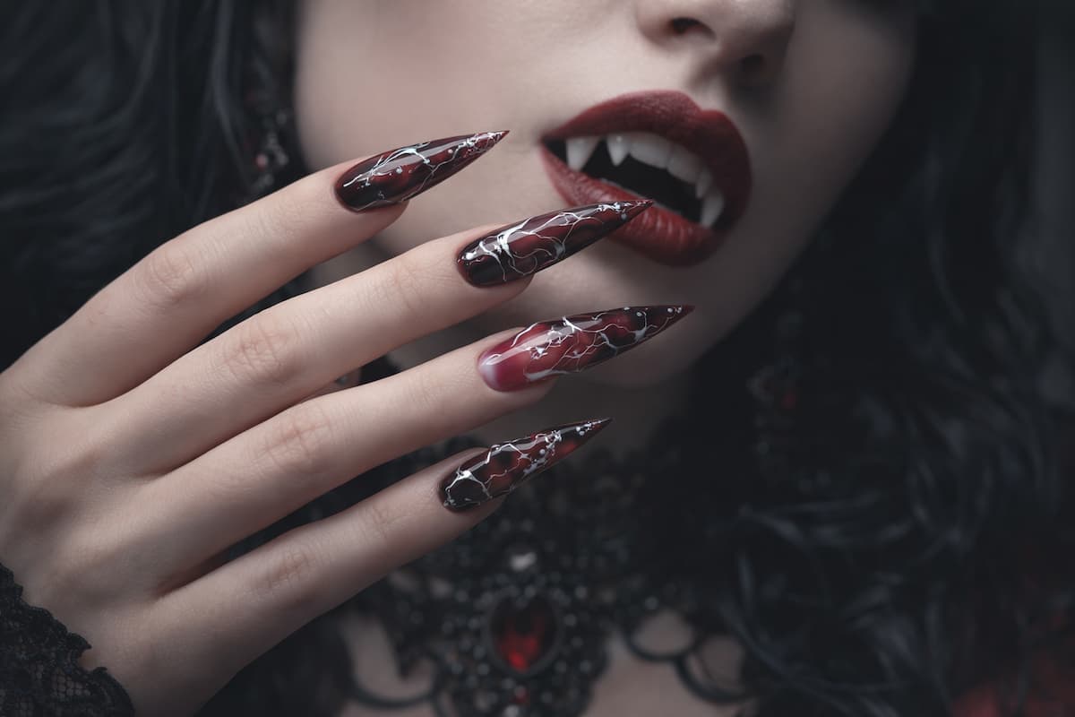 Close-up of black and red gothic stiletto nails with marbled nail art and claw shape, worn with dark lipstick and dramatic goth makeup.