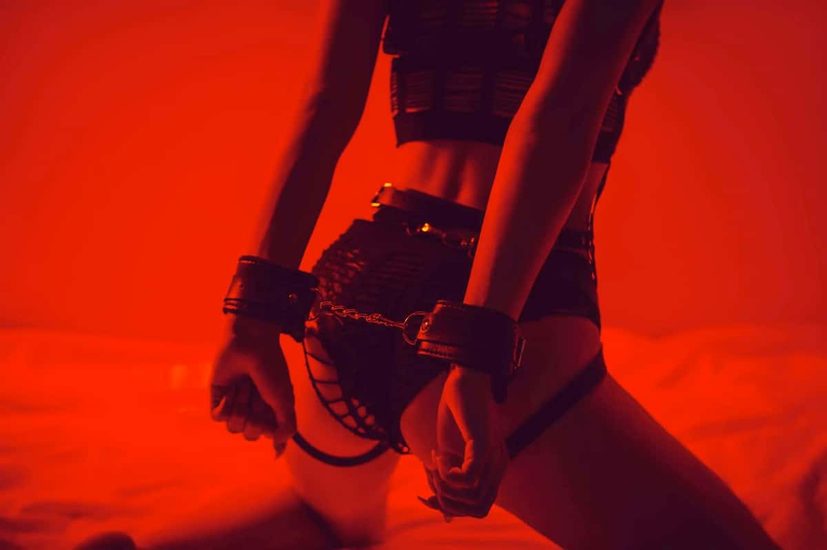 Person kneeling on a bed in red lighting, wearing black lingerie and leather wrist cuffs connected by a chain, illustrating beginner BDSM restraint play.