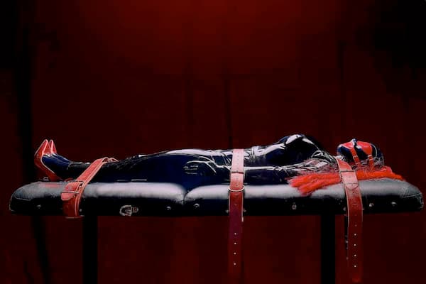 Beatrix Devine entirely covered in black latex, strapped down to a leather bondage table by red leather straps.