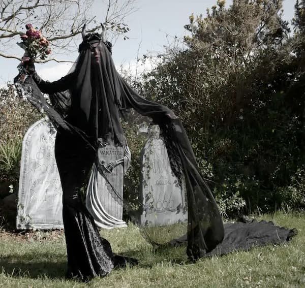 Gothic bride in black veil and crown poses in graveyard wearing MoonMaidenClothing veil and romantic goth bridal hair accessories.