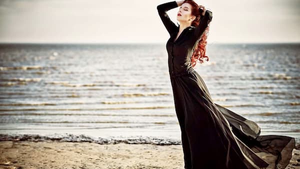 Goth woman standing on a beach with her hands in her hair, wearing a long black flowing dress and gazing toward the horizon as waves ripple in the background.