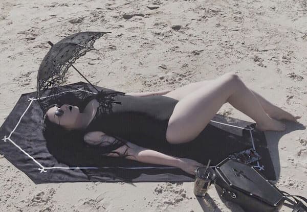Goth woman sunbathing on a beach, wearing a black swimsuit and holding a lace parasol, lying on a black coffin towel with accessories.