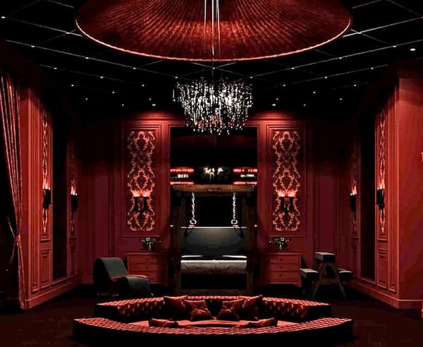 Goth sex dungeon design complete with an ornate black chandelier, red velvet couches and curtaints, plush red walls, and liberator BDSM furniture.