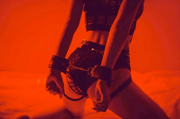Person kneeling on a bed in red lighting, wearing black lingerie and leather wrist cuffs connected by a chain, illustrating beginner BDSM restraint play.