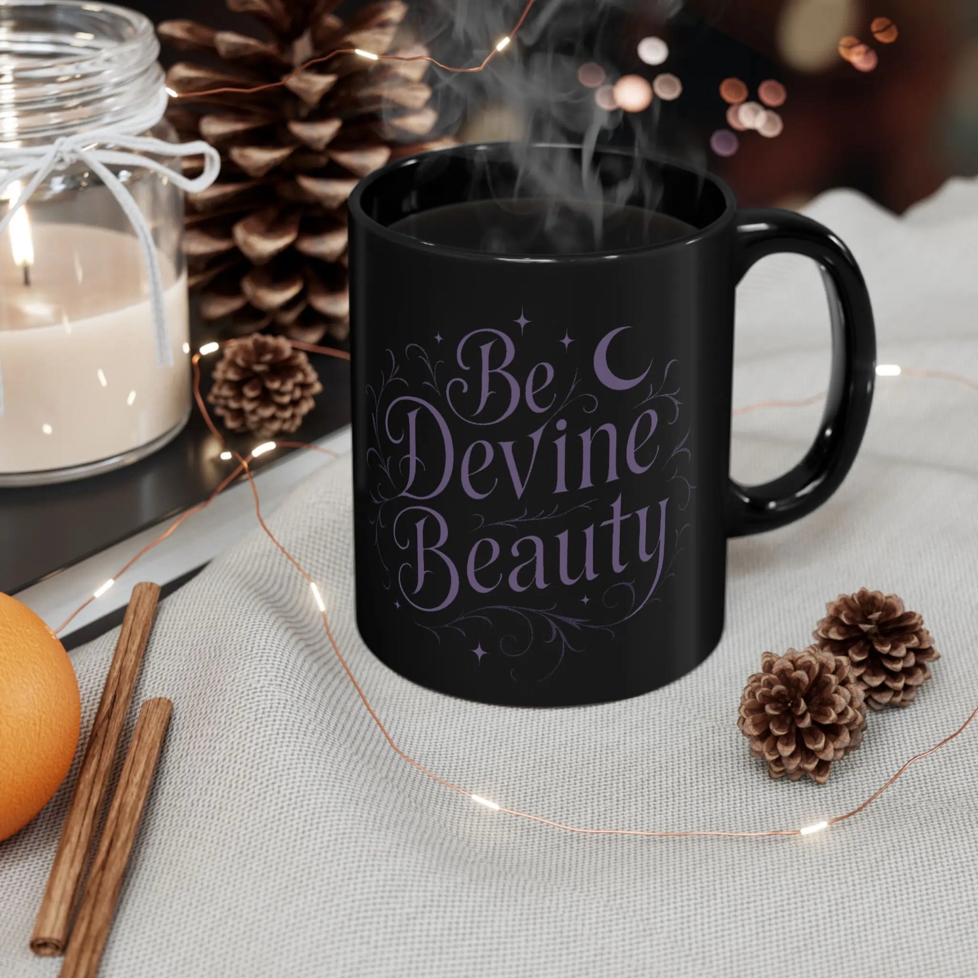 Black mug with 'Be Devine Beauty' text on a table with candles, pinecones, and lights.